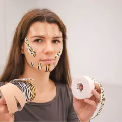 Kobieta z taśmami na twarzy, przygotowująca się do zabiegu taping twarzy. W rękach trzyma rolki taśm.