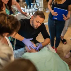 Mężczyzna w rękawiczkach wykonuje masaż kobido na twarzy kobiety. Wokół zgromadzone osoby obserwują pokaz.