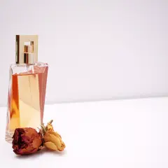 Elegancka butelka perfum z płynem w kolorze bursztynu i złotą nakrętką, obok dwie zasuszone róże. Inspiracja do tworzenia własnych perfum.