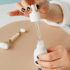Kwas glikolowy i niacynamid w kroplach serum. Dłoń z ciemnym lakierem do paznokci trzyma pipetę nad białą buteleczką.
