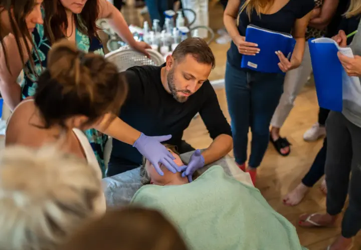 Mężczyzna w rękawiczkach wykonuje masaż kobido na twarzy kobiety. Wokół zgromadzone osoby obserwują pokaz.