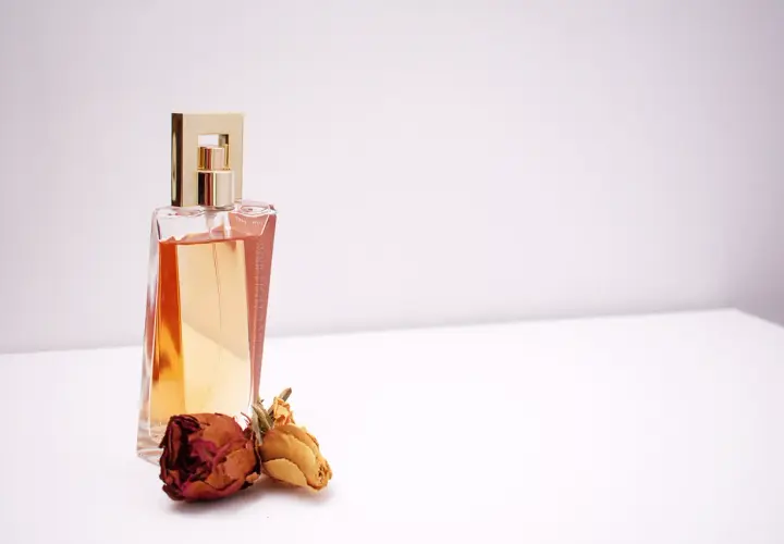 Elegancka butelka perfum z płynem w kolorze bursztynu i złotą nakrętką, obok dwie zasuszone róże. Inspiracja do tworzenia własnych perfum.