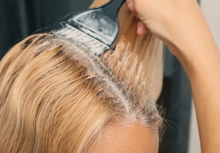 Dłoń z pędzlem nakłada rozjaśniacz na blond włosy. Prosty sposób, jak rozjaśnić włosy w domu.
