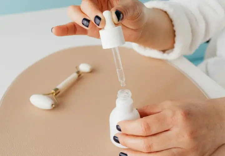 Kwas glikolowy i niacynamid w kroplach serum. Dłoń z ciemnym lakierem do paznokci trzyma pipetę nad białą buteleczką.