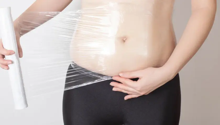Osoba owija się folią spożywczą, przygotowując się do zabiegu body wrapping.