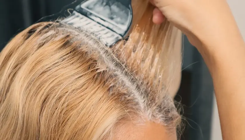 Dłoń z pędzlem nakłada rozjaśniacz na blond włosy. Prosty sposób, jak rozjaśnić włosy w domu.