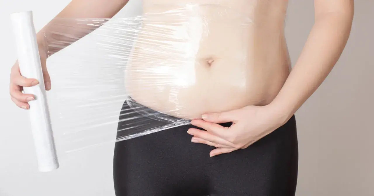 Osoba owija się folią spożywczą, przygotowując się do zabiegu body wrapping.