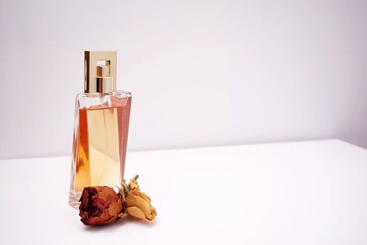 Elegancka butelka perfum z płynem w kolorze bursztynu i złotą nakrętką, obok dwie zasuszone róże. Inspiracja do tworzenia własnych perfum.