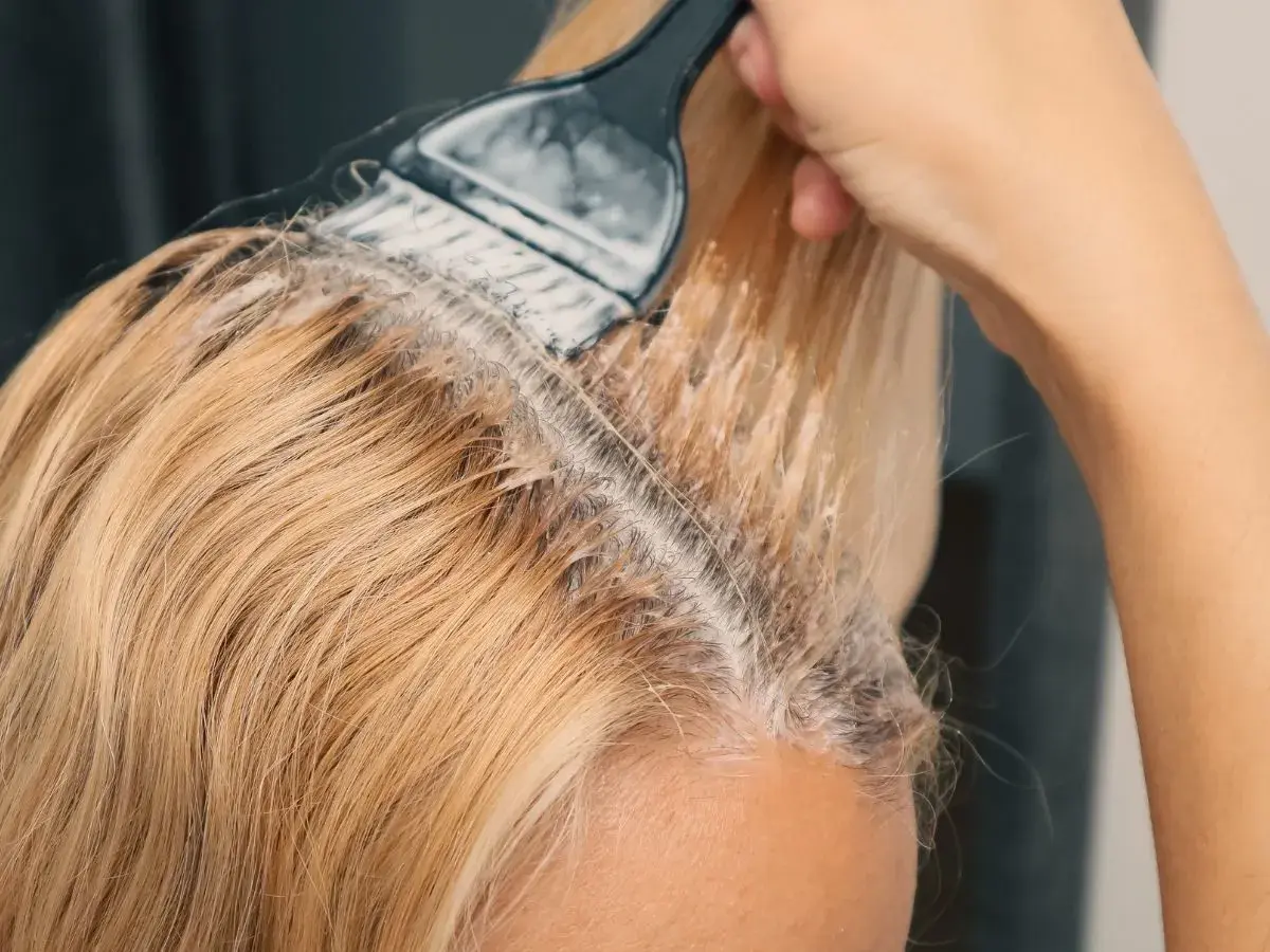 Dłoń z pędzlem nakłada rozjaśniacz na blond włosy. Prosty sposób, jak rozjaśnić włosy w domu.