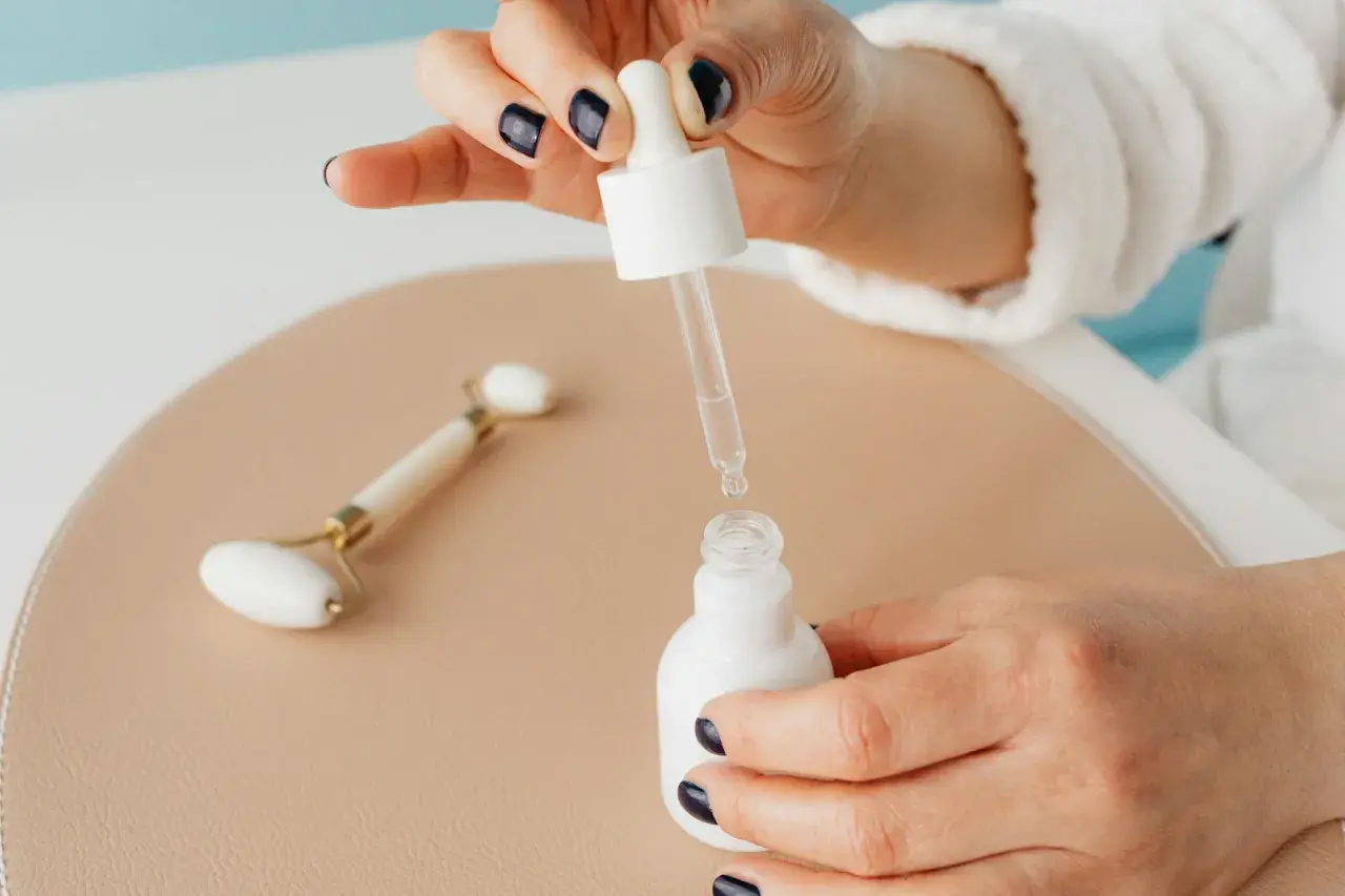 Kwas glikolowy i niacynamid w kroplach serum. Dłoń z ciemnym lakierem do paznokci trzyma pipetę nad białą buteleczką.