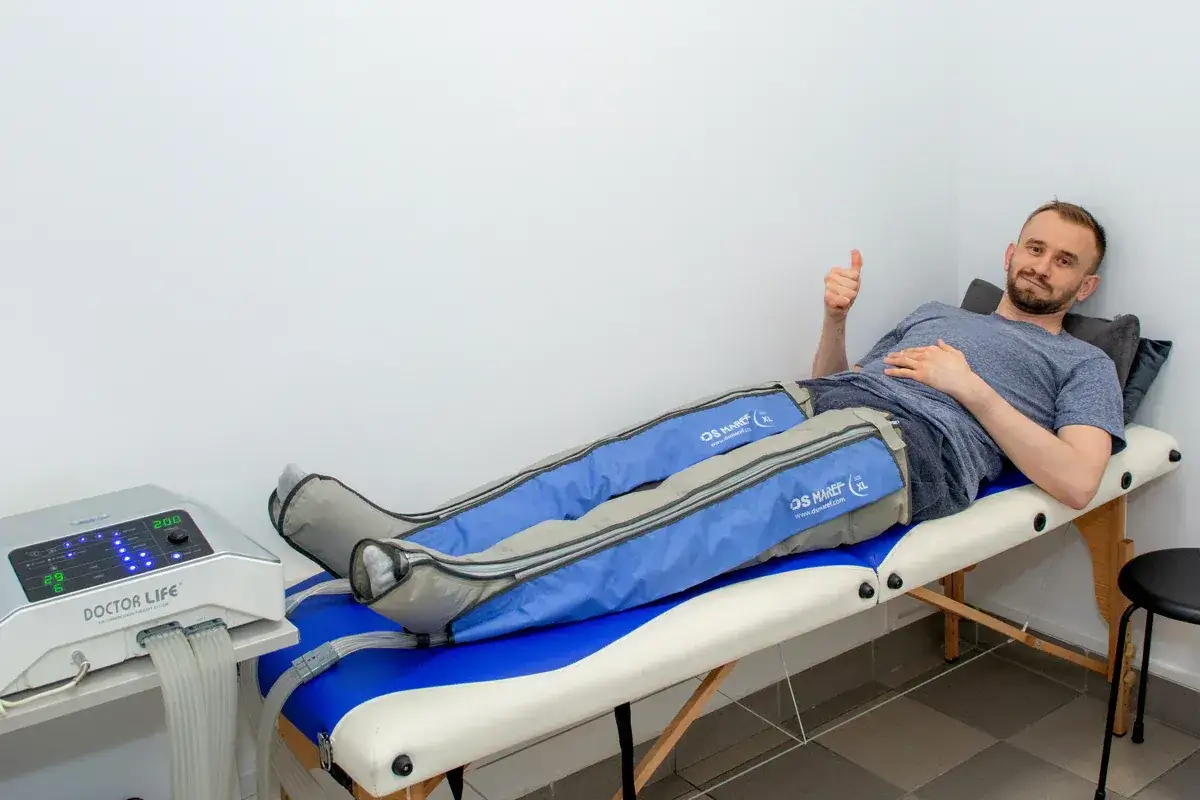 Mężczyzna leży na kozetce, poddając się zabiegowi drenażu limfatycznego nóg. Pokazuje kciuk w górę, wyrażając zadowolenie.