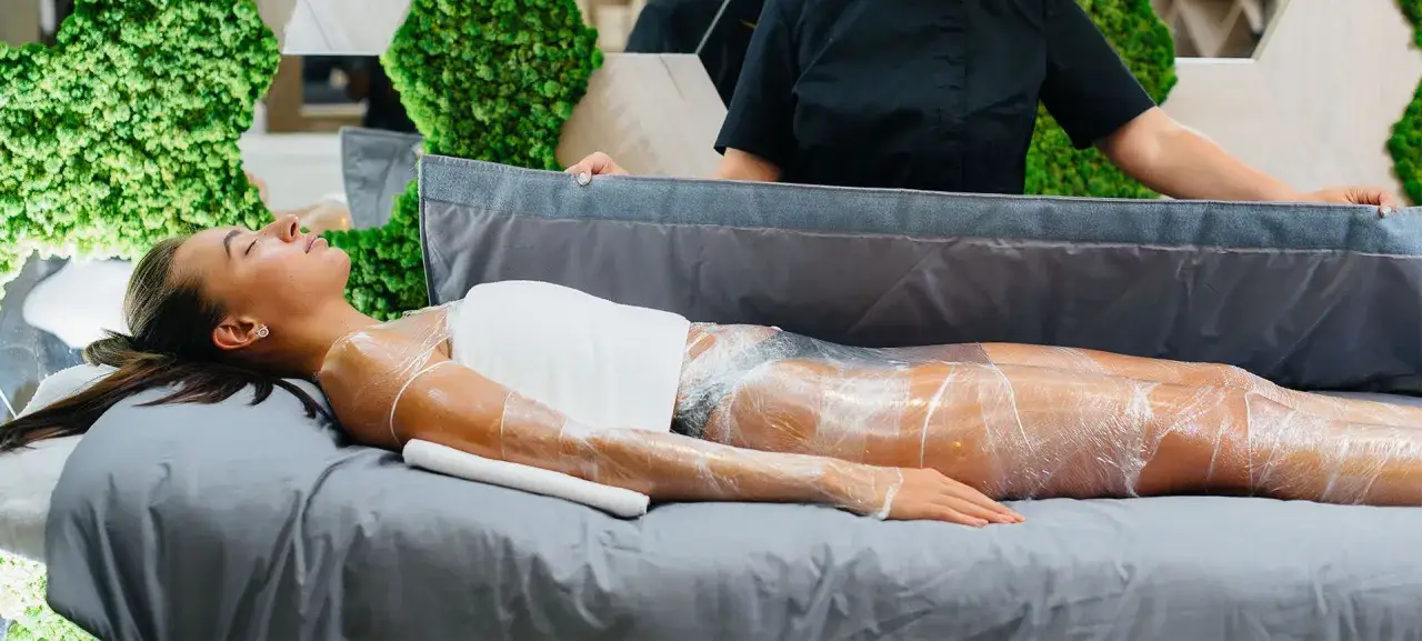 Kobieta leży na ł&oacute;żku, owinięta folią podczas zabiegu body wrapping. W tle zielone dekoracje.