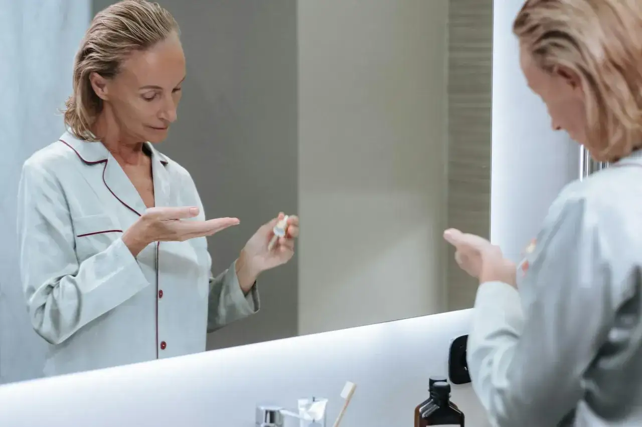 Kobieta w piżamie aplikuje serum z retinolem na trądzik do dłoni, patrząc w lustro.